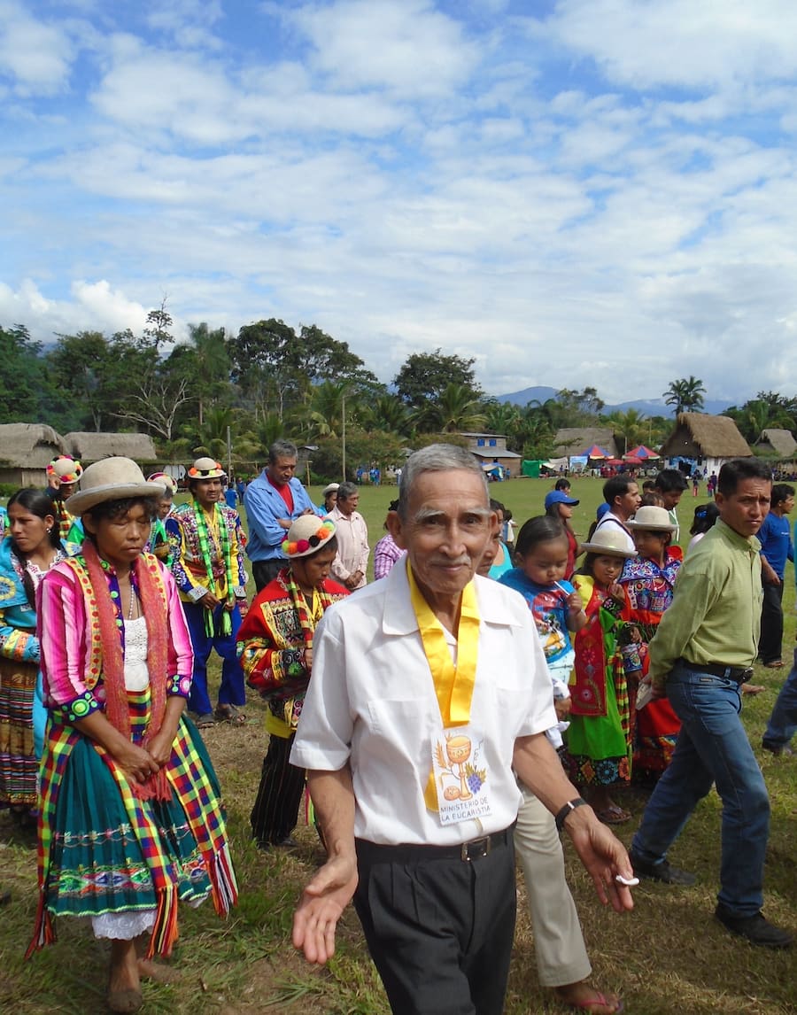 Procession of locals in a field surrounded by thatched buildings.