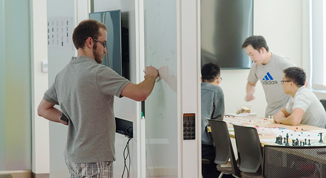 Students in the Game Lab develop games: a student writes on a board and other students work at a table covered in game pieces.