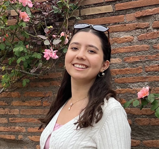 Daniela smiling in front of a red brick wall with pink flowers